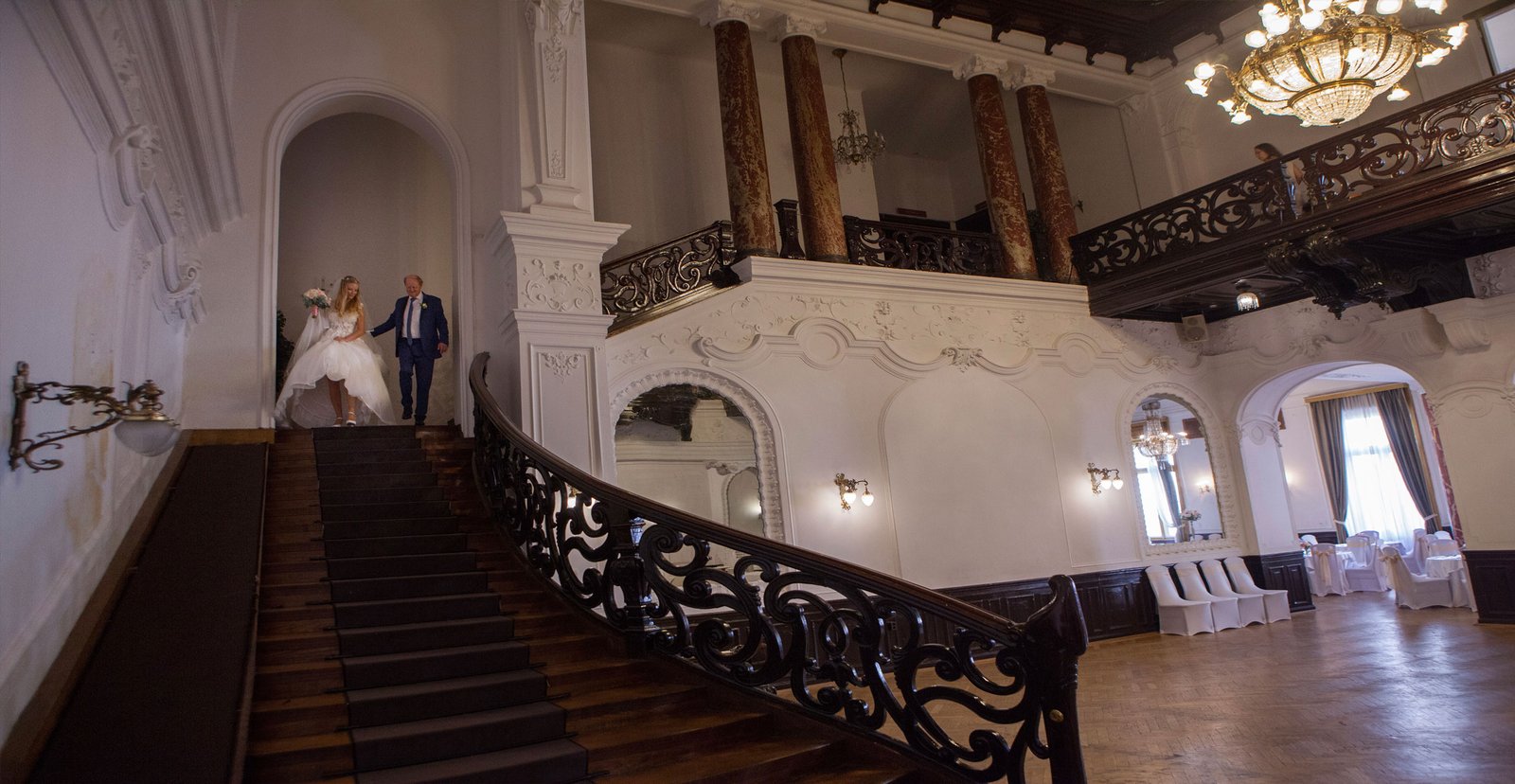 In a unique castle interior, a proud father leads the bride down a grand staircase to the wedding ceremony.